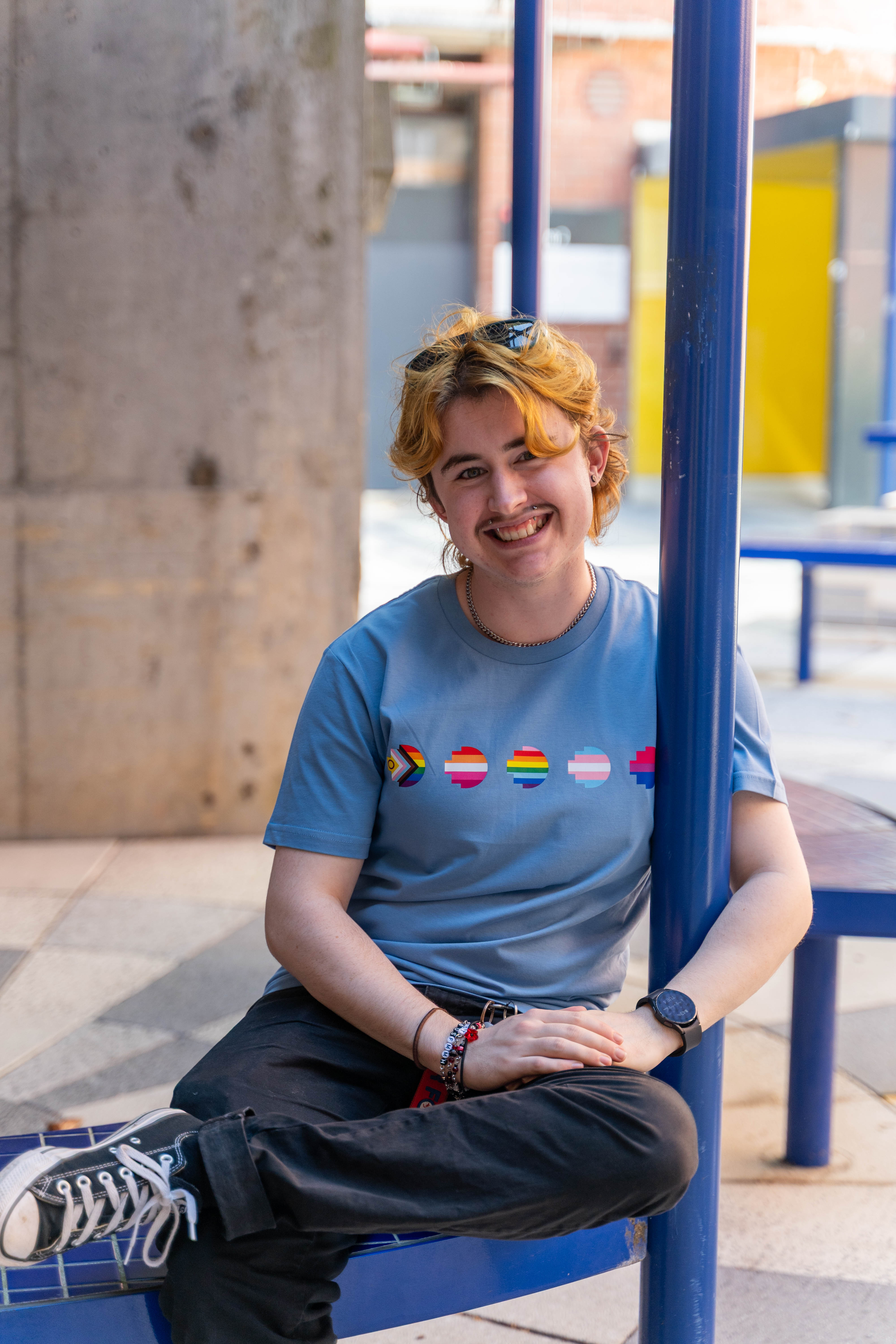 Casual pose of a person wearing the limited edition blue Pride Unity Tee from RMIT Store, seated with pride flag designs on the chest against a stylish background.